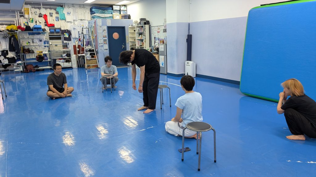 Instructor bending over to speak to a small group seated on stools and the floor in a bright blue gym studio.